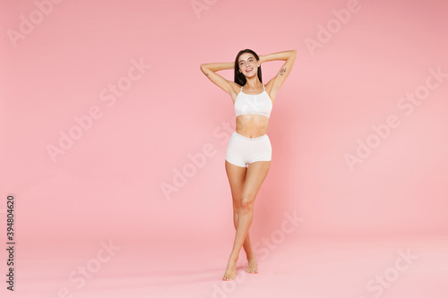 Full length of smiling cheerful attractive young brunette woman in white underwear showing fit sports body posing hold hands behind head looking aside isolated on pink background, studio portrait.