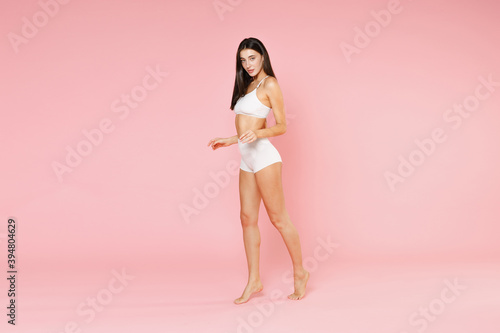 Full length side view of beautiful young brunette woman in white underwear showing fit sexy body posing standing on toes spreading hands looking camera isolated on pink background, studio portrait.