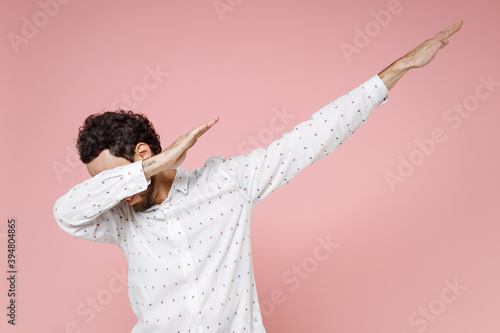 Cheerful young bearded man 20s in basic casual white shirt standing doing dab hip hop dance hands gesture, youth sign hiding and covering face isolated on pastel pink color background studio portrait.