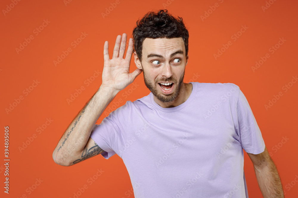 Shocked curious young bearded man 20s wearing basic casual violet t-shirt standing try to hear you overhear listening intently looking aside isolated on bright orange color background studio portrait. Shocked curious young bearded man 20s wearing basic casual violet t-shirt standing try to hear you overhear listening intently looking aside isolated on bright orange color background studio portrait.