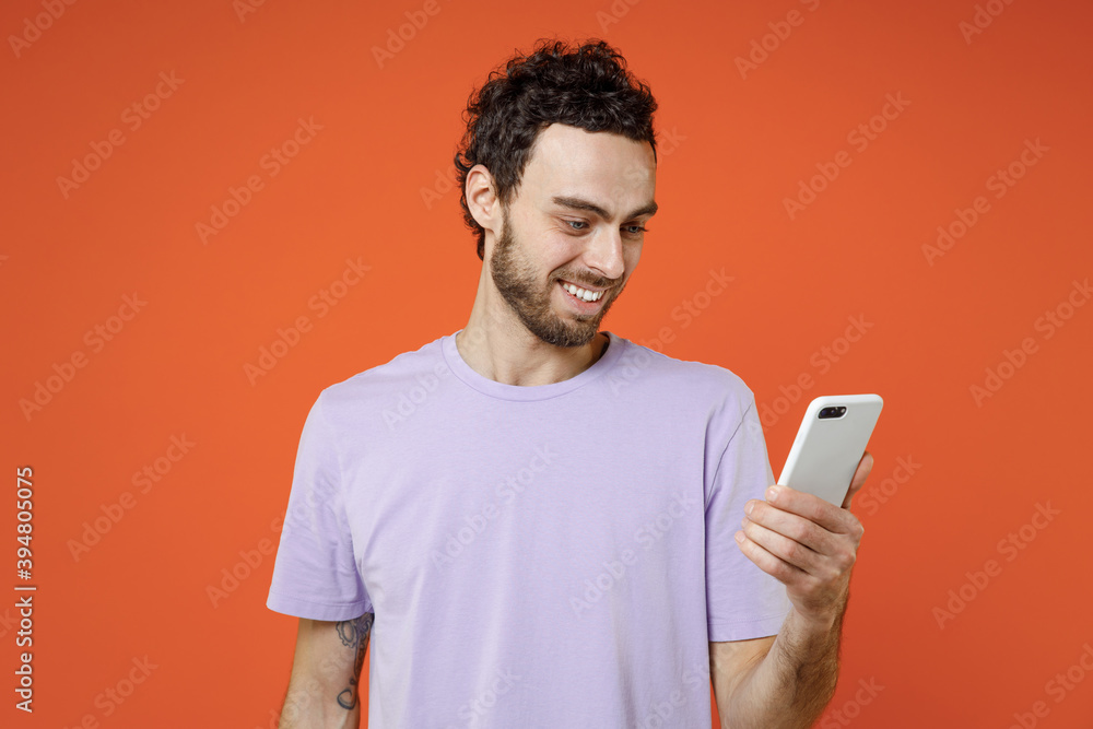 Cheerful smiling handsome attractive young bearded man 20s in casual violet t-shirt standing using mobile cell phone typing sms message isolated on bright orange color wall background studio portrait. Cheerful smiling handsome attractive young bearded man 20s in casual violet t-shirt standing using mobile cell phone typing sms message isolated on bright orange color wall background studio portrait.