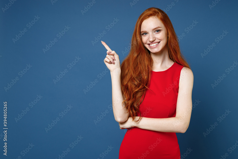 Smiling beautiful young redhead woman 20s wearing bright red elegant evening dress standing pointing index finger aside up on mock up copy space isolated on blue color wall background studio portrait. Smiling beautiful young redhead woman 20s wearing bright red elegant evening dress standing pointing index finger aside up on mock up copy space isolated on blue color wall background studio portrait.
