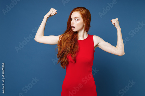 Shocked amazed young redhead woman 20s wearing bright red elegant evening dress standing spreading hands showing biceps muscles looking aside isolated on blue color wall background studio portrait.