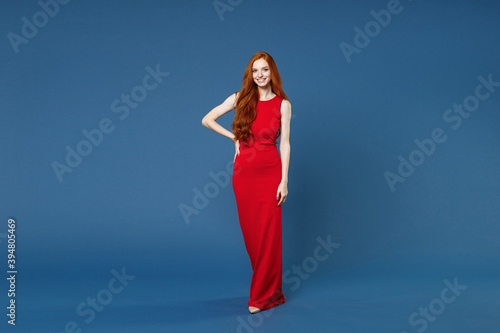 Full length of smiling pretty stunning beautiful young redhead woman 20s wearing bright red elegant evening dress standing and looking camera isolated on blue color wall background studio portrait.