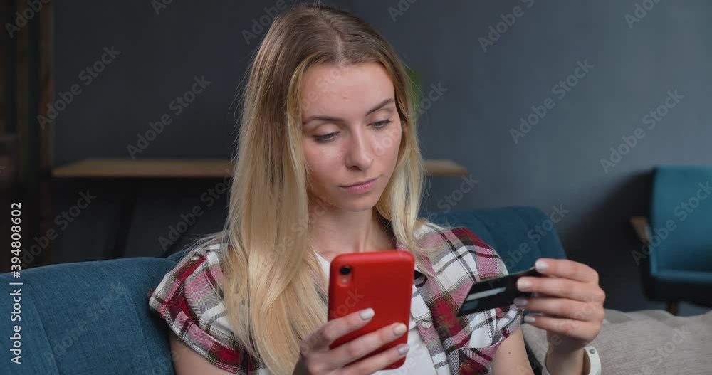 Young caucasian Blonde Woman is relaxing at Home, shopping online with Credit Card from Home. The Female is sitting at Home, surfing Online Shops on her Smartphone. Orders Home Delivery. Technology.