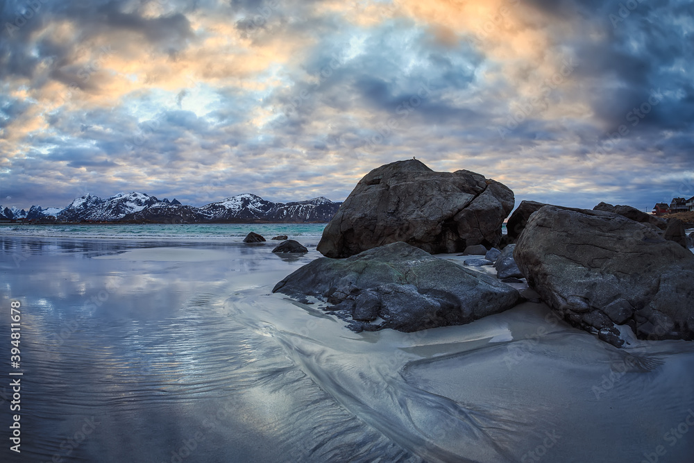 Obraz premium rocks in the sand of the beach wet by sea water with mountains in the background during a cloudy day with rainbow colors above the clouds