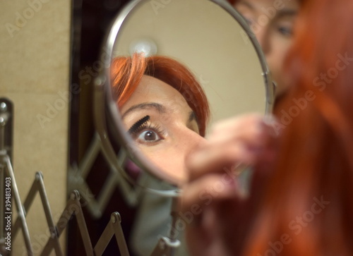 Young white woman with red hair makes up in front of the mirror with the brush on her eyelashes, three mirrors