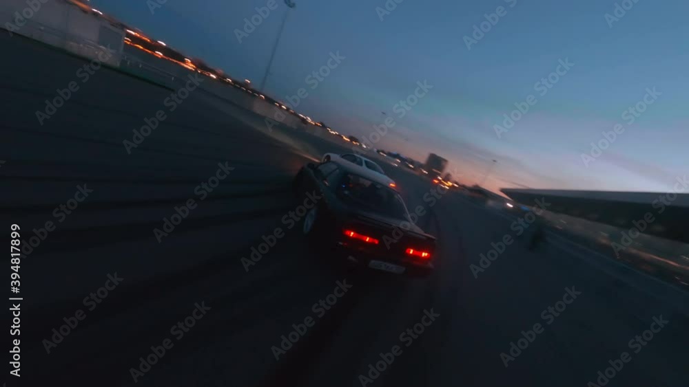 Fast sports jdm cars drift follow race driving about special ground near modern building against illuminated city in evening first point view fpv sport drone cinematic shot.