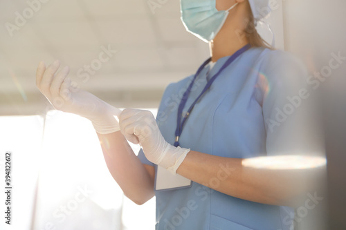 Doctor in protective mask and scrubs putting on medical gloves indoors, closeup