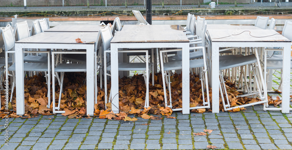 Useless restaurant, pub or beer garden chairs and tables folded and tied because of coronavirus lockdown. Old autumn leaves under empty gastronomy seats. Pandemic problems of small business.