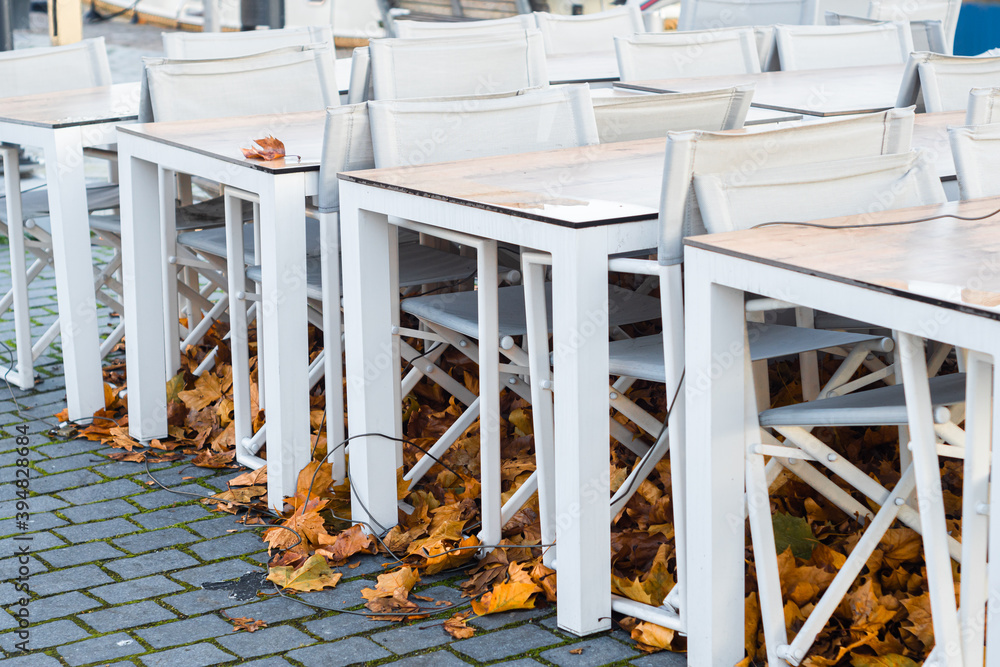 Useless restaurant, pub or beer garden chairs and tables folded and tied because of coronavirus lockdown. Old autumn leaves under empty gastronomy seats. Pandemic problems of small business.