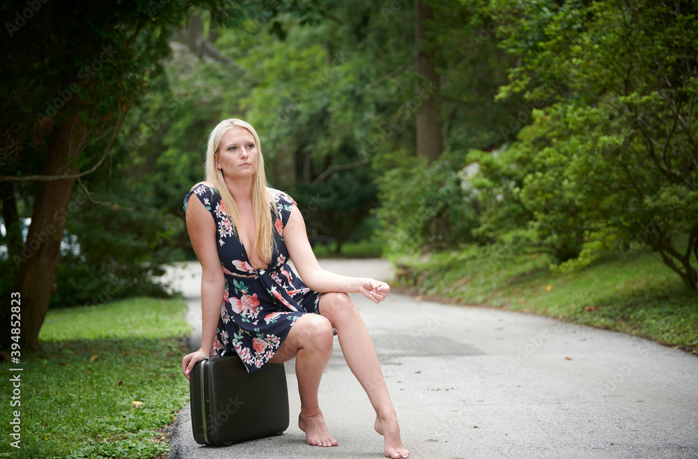 Sexy young blonde woman poses along country road wearing sundress or romper with plunging neckline - sitting on suitcase