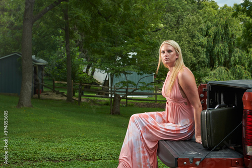 Sexy young blonde woman poses on or near farm, sitting on truck tailgate, wearing long flowing pink dress - with suitcase nearby - cancelled trip theme