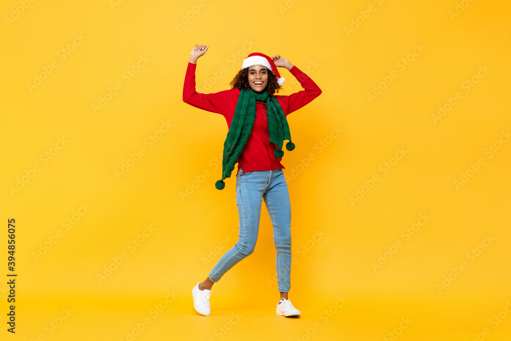 Fototapeta premium Fun portrait of happy African American woman in Christmas attire smiling and raising hands on isolated yellow studio background