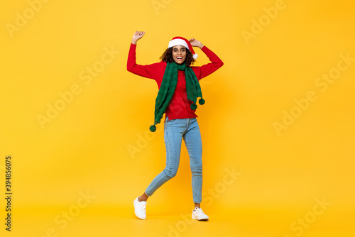 Fun portrait of happy African American woman in Christmas attire smiling and raising hands on isolated yellow studio background © Atstock Productions