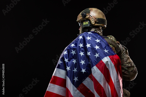 Back view of a military man in a hard hat and full gear, draped over the shoulder of flag of America isolated on black. The concept of patriotism, protection of the Motherland, love for the country