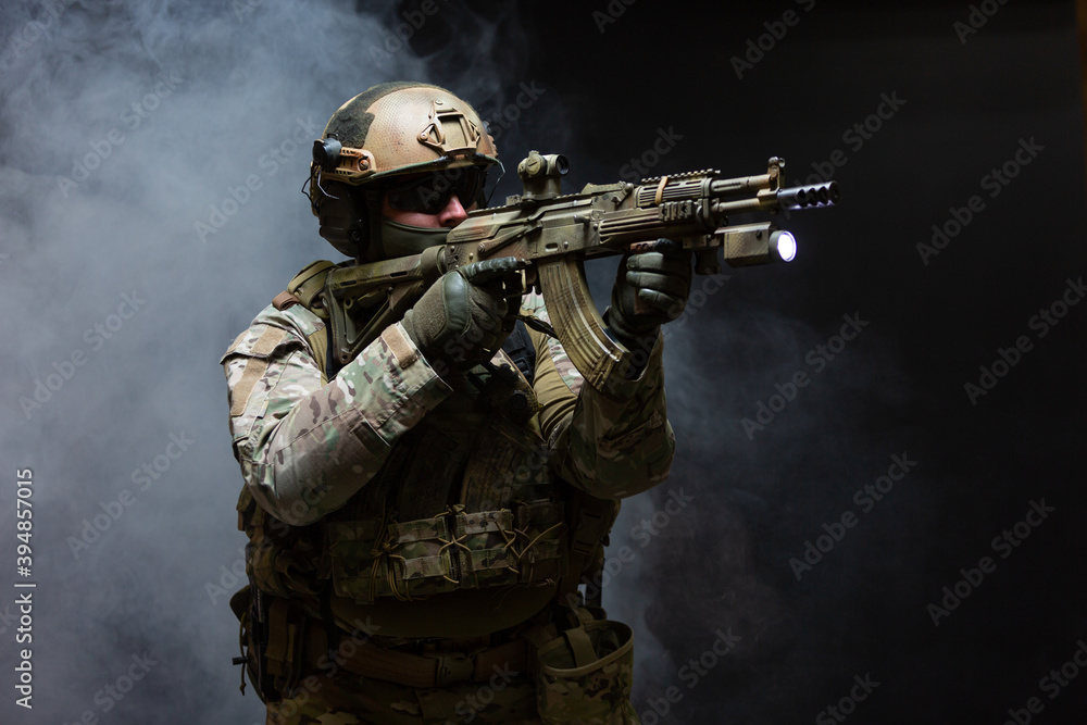 Side view of army soldier in military camouflage uniform, helmet, glasses and mask holds rifle near face and takes aim using collimator sight standing in attack position, in smoke on black background Side view of army soldier in military camouflage uniform, helmet, glasses and mask holds rifle near face and takes aim using collimator sight standing in attack position, in smoke on black background
