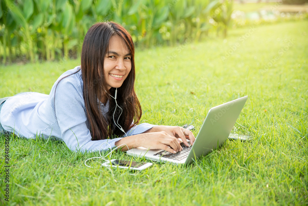 Working online from Home Concept. Asian Thai girl typing laptop and review annual report in business market.Woman lying on green grass and use computer via internet to send data and search on digital. Working online from Home Concept. Asian Thai girl typing laptop and review annual report in business market.Woman lying on green grass and use computer via internet to send data and search on digital.