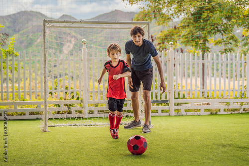 Little cute kid boy in red football uniform and his trainer or father playing soccer  football on field  outdoors. Active child making sports with kids or father  Smiling happy boy having fun in