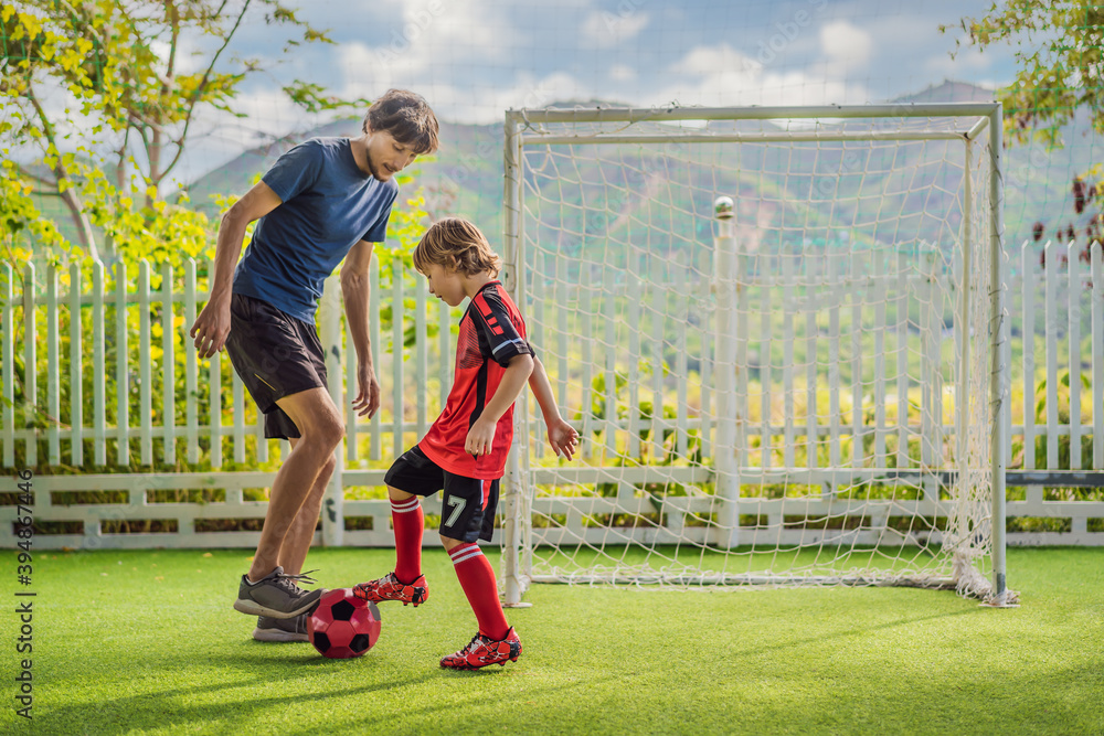 Obraz premium Little cute kid boy in red football uniform and his trainer or father playing soccer, football on field, outdoors. Active child making sports with kids or father, Smiling happy boy having fun in