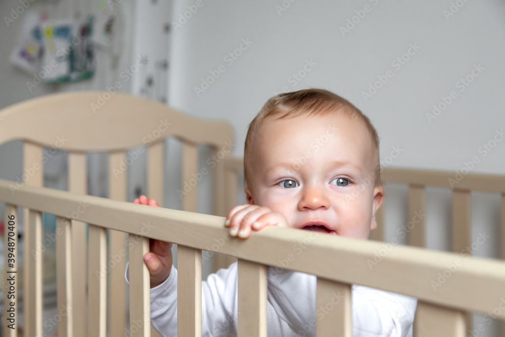 Blond cute llittle kid in white bodysuit stands in wooden bed Blond cute llittle kid in white bodysuit stands in wooden bed