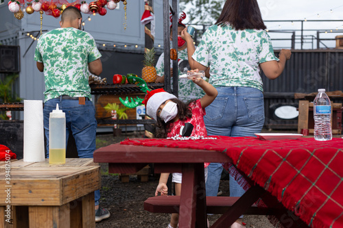 Latin girl with a mask dressed a red tie dye sweater helps prepare Christmas dinner is a barbecue of fresh vegetables and meats in a food truck place. Christmas family dinner concept in the new normal