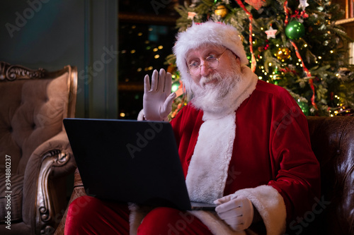 Christmas online congratulations from Santa. Santa Claus using notebook laptop for distance Christmas call kids sitting near Christmas tree at home. Happy New Year Covid 2021.