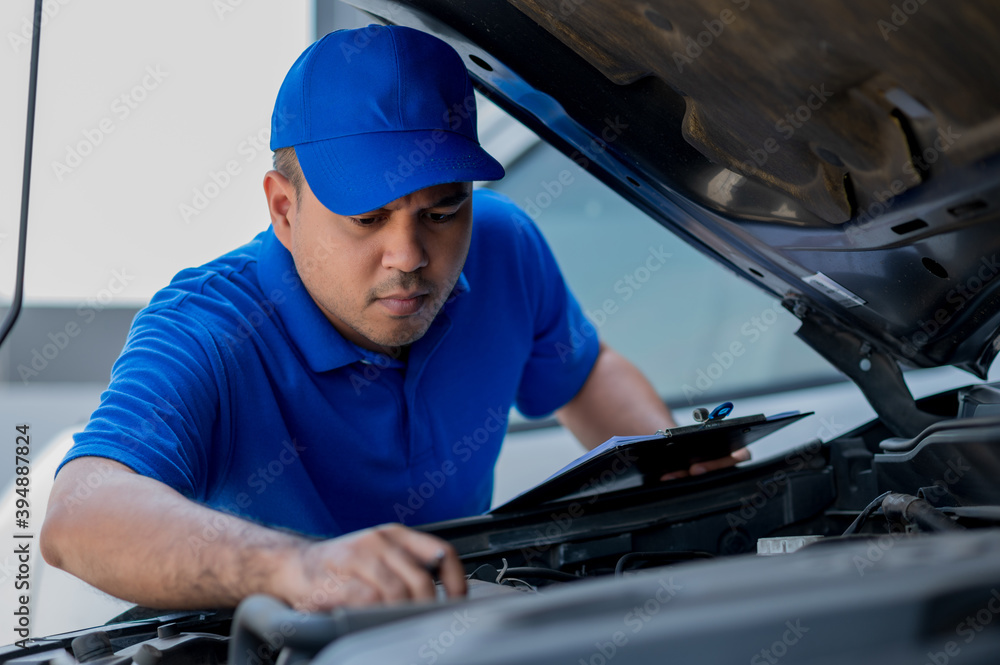 A young Asian auto mechanic opens the bonnet. To check for engine damage And perform professional maintenance. He wearing blue uniform. A young Asian auto mechanic opens the bonnet. To check for engine damage And perform professional maintenance. He wearing blue uniform.