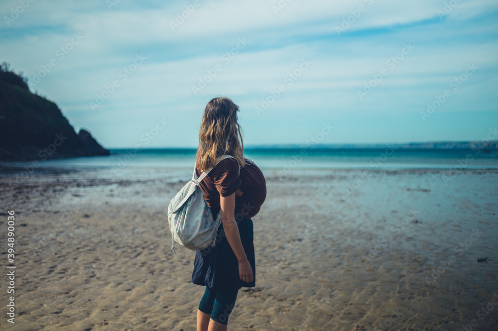 Young mother with her baby in a sling is standing on the beach