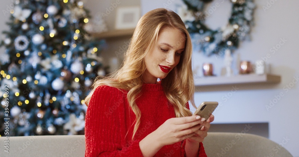 Close up portrait of joyful young beautiful Caucasian woman in decorated modern room with xmas tree texting on smartphone and sending greetings on Christmas Eve. Festive mood. Xmas spirit concept Close up portrait of joyful young beautiful Caucasian woman in decorated modern room with xmas tree texting on smartphone and sending greetings on Christmas Eve. Festive mood. Xmas spirit concept