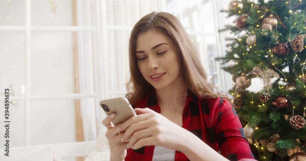 Close up portrait of joyful young beautiful Caucasian woman in room with christmas tree tapping and texting on smartphone wishing congratulation on Christmas Eve. Holidays concept Close up portrait of joyful young beautiful Caucasian woman in room with christmas tree tapping and texting on smartphone wishing congratulation on Christmas Eve. Holidays concept
