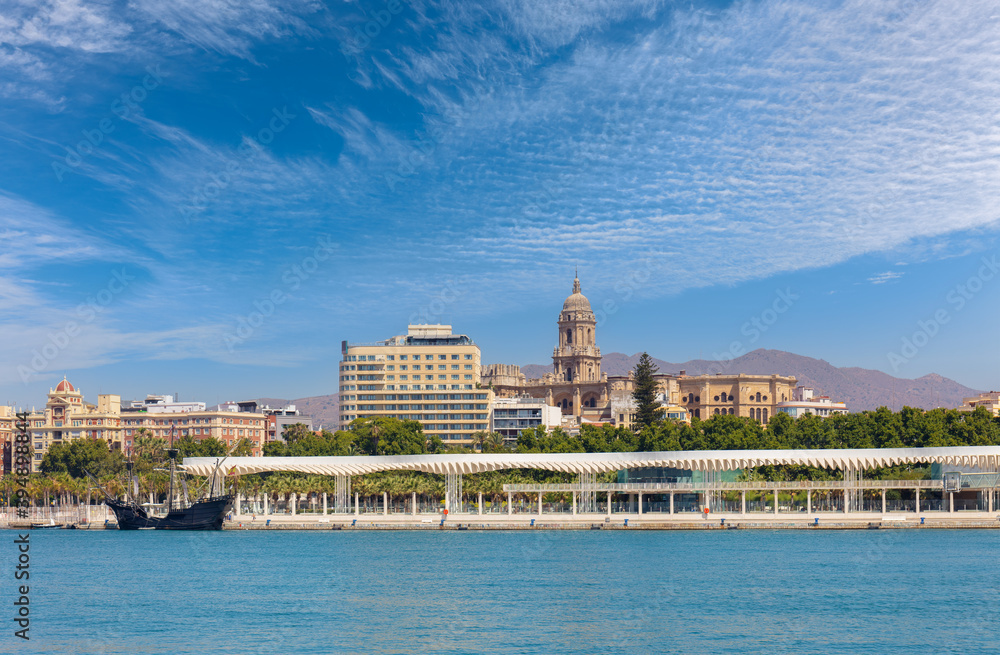 View of the port and skyline of the Spanish port city of Malaga in Andalusia. It's a summer day with blue skies. On the left is a historic sailing ship. In the background are mountains.