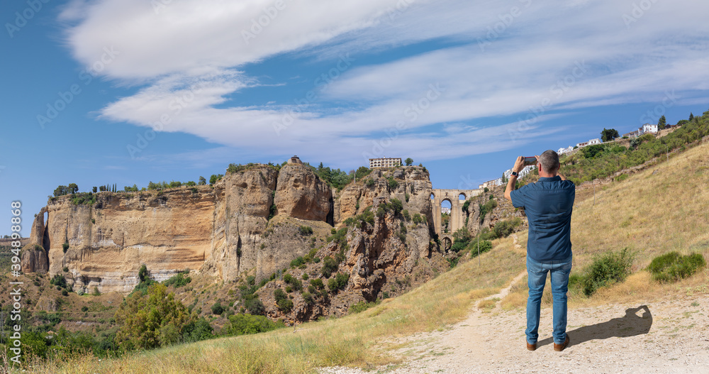 Naklejka premium A male tourist photographs the famous arched bridge of Ronda in Andalusia in summer with a cloudy blue sky. The city is on top of the plateau. At the very bottom is a small waterfall.