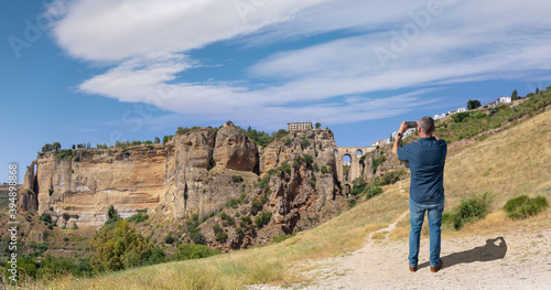 A male tourist photographs the famous arched bridge of Ronda in Andalusia in summer with a cloudy blue sky. The city is on top of the plateau. At the very bottom is a small waterfall.