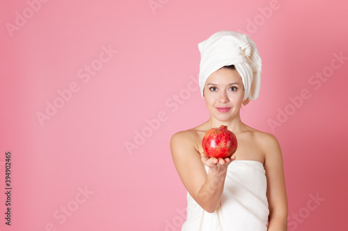 mock up, portrait of a beautiful young woman with a towel on her head and on her body holding out a pomegranate to us, isolated on a pink background.