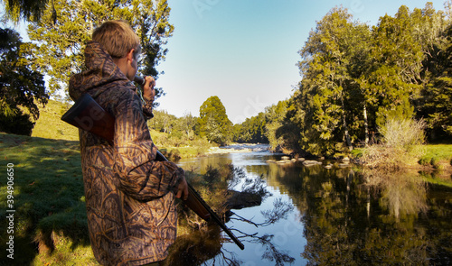 Boy dressed in camouflage clothes holding shot gun and calling Ducks with caller on riverbank photo