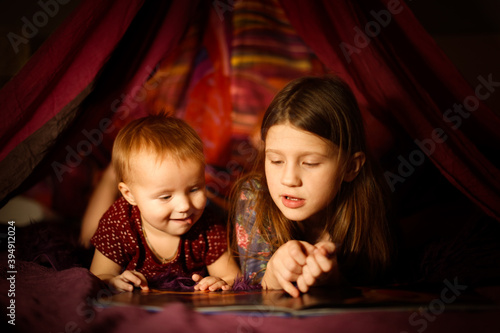 Caucasian girls children sisters read a book in a toy tent with garlands  children s games and houses  the older sister reads to the younger  dark cozy photos 