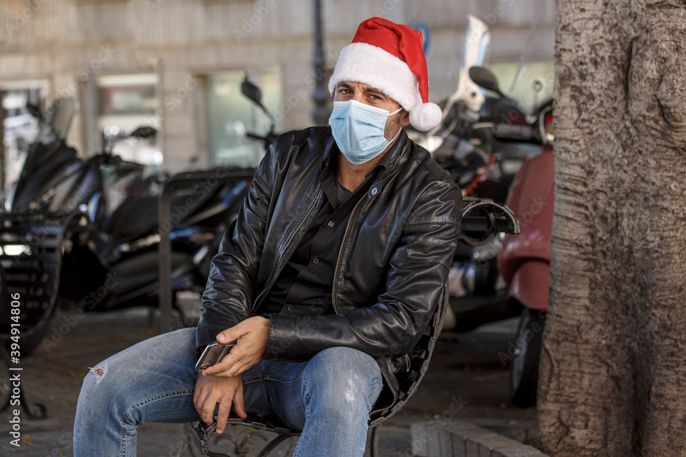 Uomo con cappello da babbo natale, giacca in pelle e mascherina chirurgica è seduto in un panchina in contesto urbano Uomo con cappello da babbo natale, giacca in pelle e mascherina chirurgica è seduto in un panchina in contesto urbano