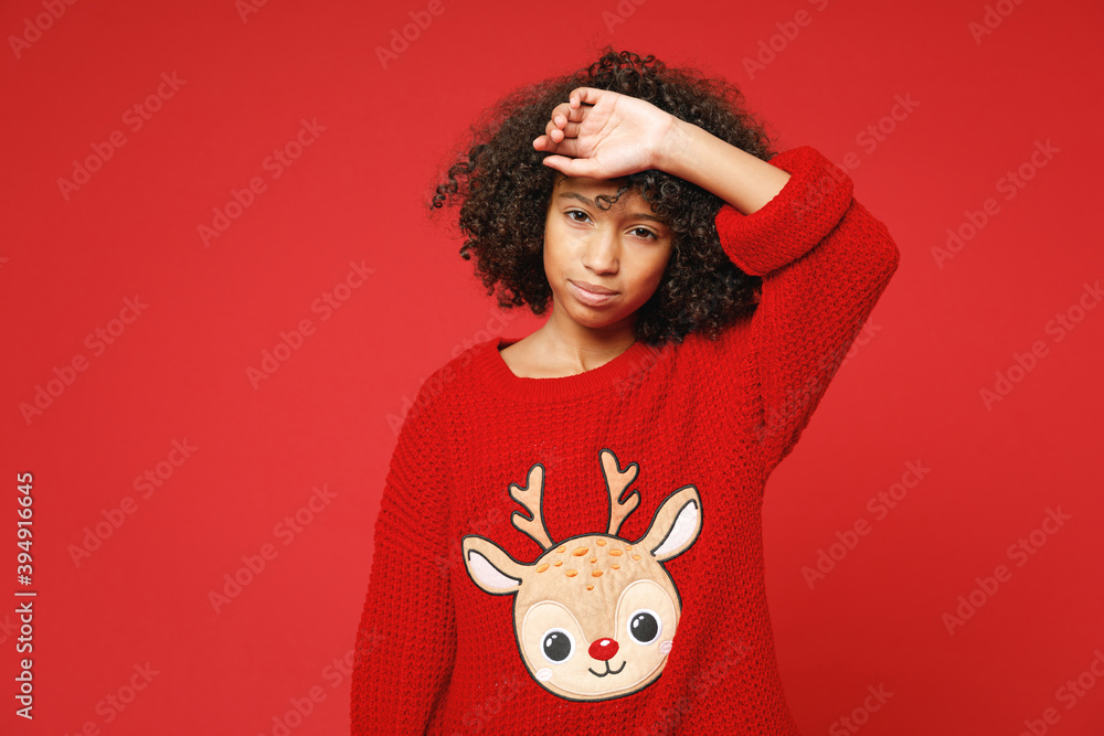 Little sad upset frustrated african american curly kid girl 12-13 years old wear knitted deer Christmas sweater isolated on bright red background children studio portrait. Childhood lifestyle concept. Little sad upset frustrated african american curly kid girl 12-13 years old wear knitted deer Christmas sweater isolated on bright red background children studio portrait. Childhood lifestyle concept.