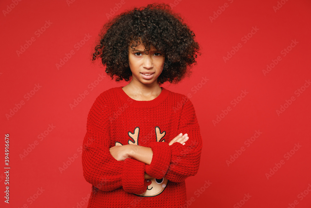 Little sad upset frustrated african american curly kid girl 12-13 years old wear knitted deer Christmas sweater isolated on bright red background children studio portrait. Childhood lifestyle concept. Little sad upset frustrated african american curly kid girl 12-13 years old wear knitted deer Christmas sweater isolated on bright red background children studio portrait. Childhood lifestyle concept.
