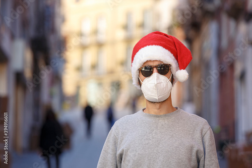 Uomo con cappello da babbo natale, maglia grigia e mascherina ffp2 in centro città tra le vetrine dei negozi photo