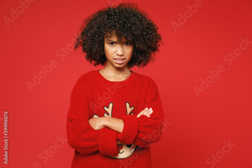 Little sad upset frustrated african american curly kid girl 12-13 years old wear knitted deer Christmas sweater isolated on bright red background children studio portrait. Childhood lifestyle concept.