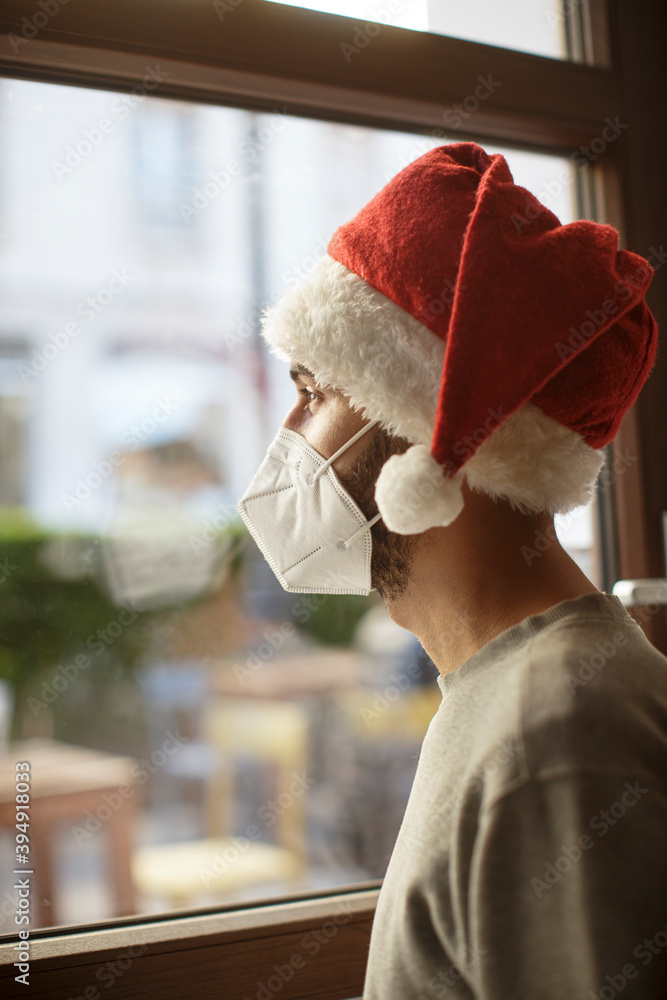 Ragazzo con cappello da babbo natale e mascherina ffp2 guarda triste fuori  casa attraverso il vetro di una finestra Stock Photo | Adobe Stock