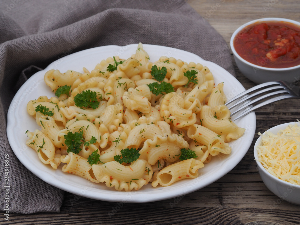 Curly pasta, macaroni with spices and herbs in a plate, tomato sauce, grated parmesan cheese, fork on a wooden table, closeup. Simple delicious rustic dish with pasta products