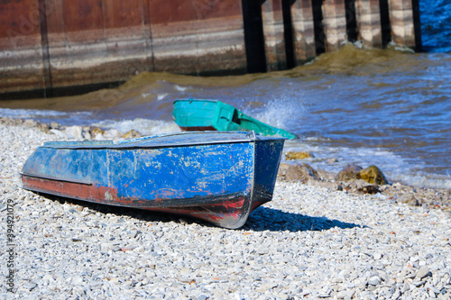 old rusty fishing boat lies on  pebble coast of the river.  waves of  tide roll over the little iron man and break into spray and foam on the side, bow and stern of the boat.