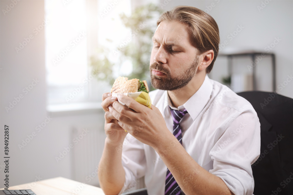 Handsome businessman in formal clothing enjoying tasty burger at office during lunch time. Busy man sitting at desk and eating fast food.