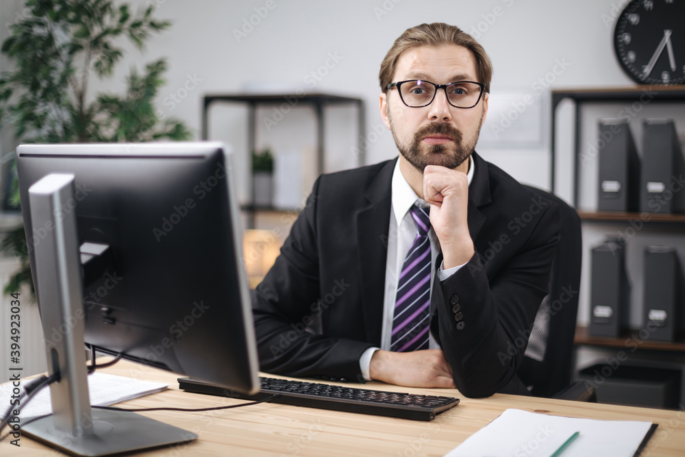 Front view of confident bearded man in black suit and eyeglasses sitting at desk with modern computer. Mature businessman enjoying favorite work at office center. Front view of confident bearded man in black suit and eyeglasses sitting at desk with modern computer. Mature businessman enjoying favorite work at office center.