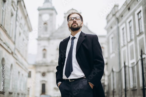 Self-sufficient bearded man in formal outfit andeyeglasses standing at old city street and holding hands in pockets. Concept of business and sucess.