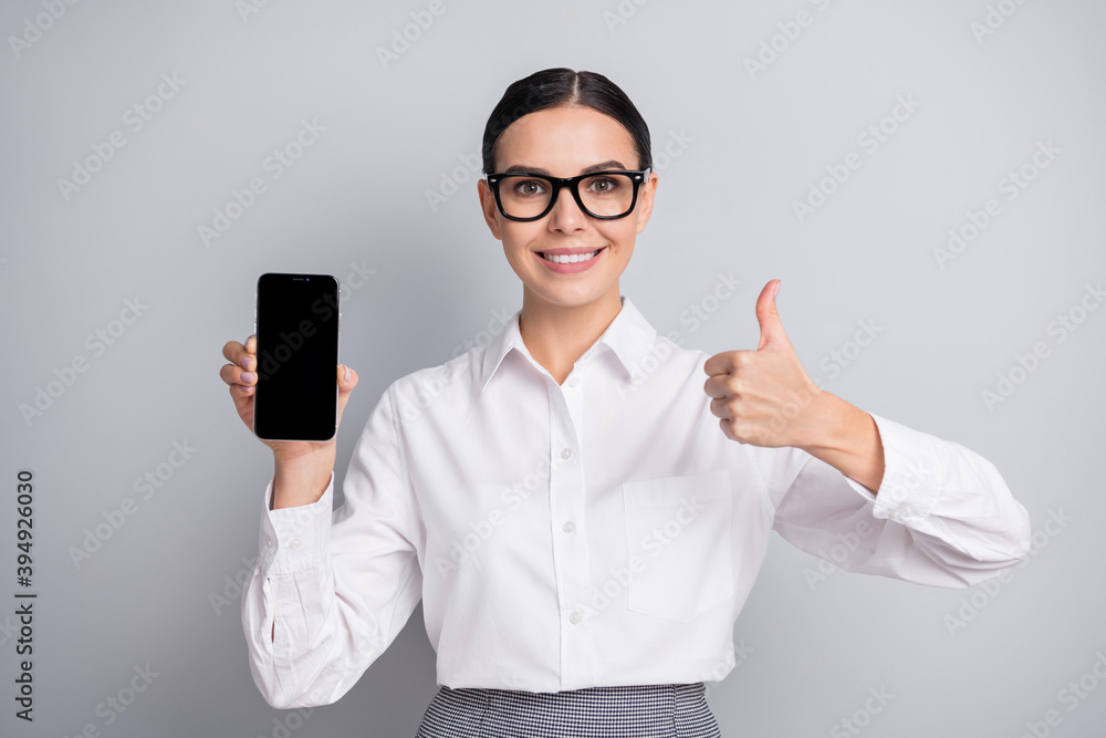 Photo of pretty promoter girl hold phone screen empty space raise thumb up wear specs white shirt isolated grey color background Photo of pretty promoter girl hold phone screen empty space raise thumb up wear specs white shirt isolated grey color background