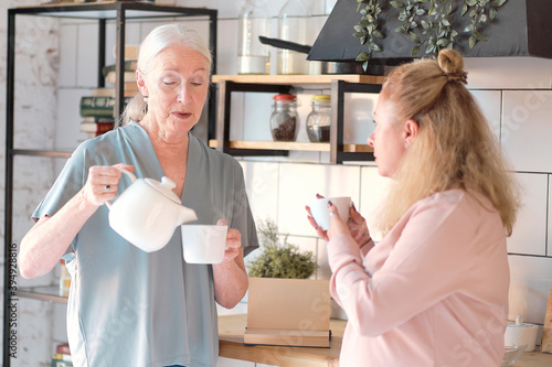 Friendly family viewing photos at home. Senior woman is enjoying a catch up with her daughter. They are drinking cups of tea in the kitchen. Elderly woman with female caregiver in living room photo